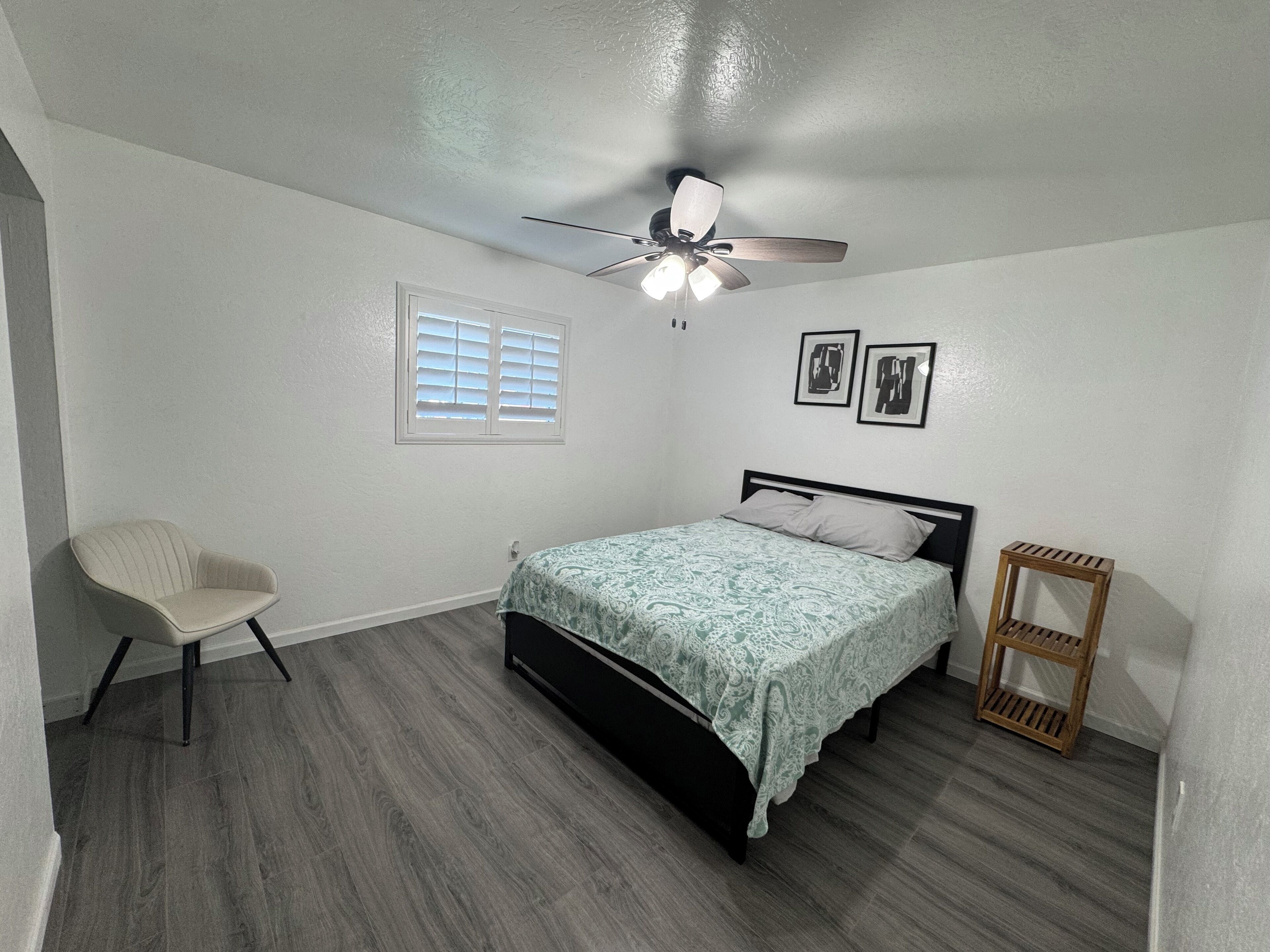 Une chambre à coucher comprenant un lit, une chaise, un ventilateur de plafond, une fenêtre et deux tableaux encadrés accrochés au mur.