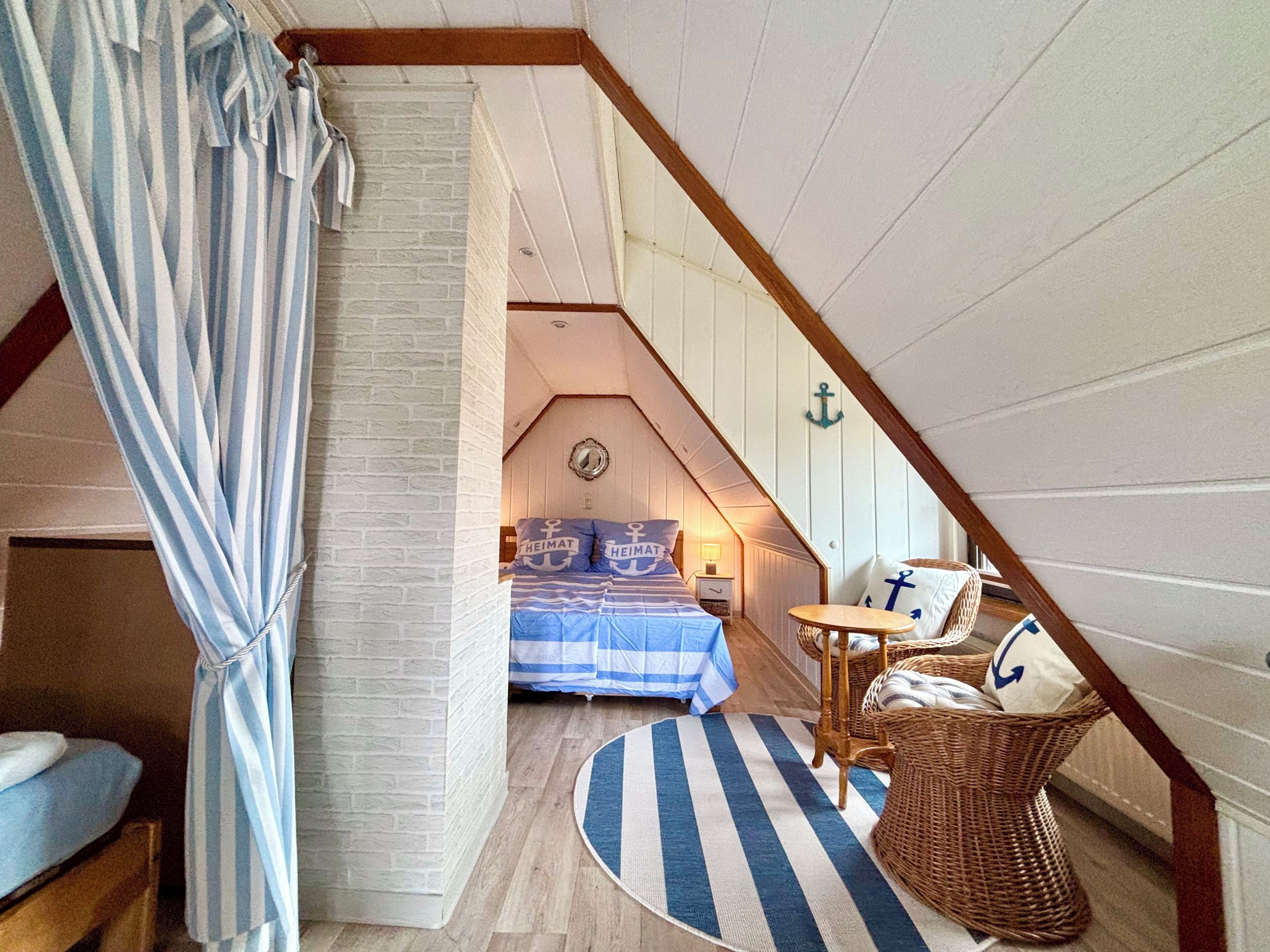 A bedroom with a sloped ceiling, a bed with nautical-themed bedding, a small round table, wicker furniture, and a striped curtain.