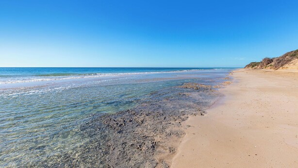 Una playa con agua cristalina, orilla arenosa y un acantilado lejano.