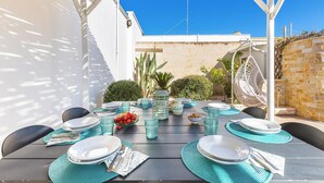 Un área de comedor al aire libre con una mesa dispuesta para cuatro personas, rodeada de sillas, bajo una pérgola.