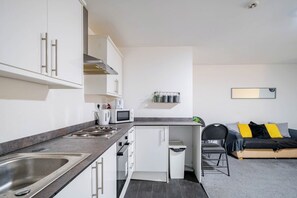Cocina compacta con lavaplatos, estufa, microondas y armarios. Hay un pequeño comedor con una silla y un sofá.
