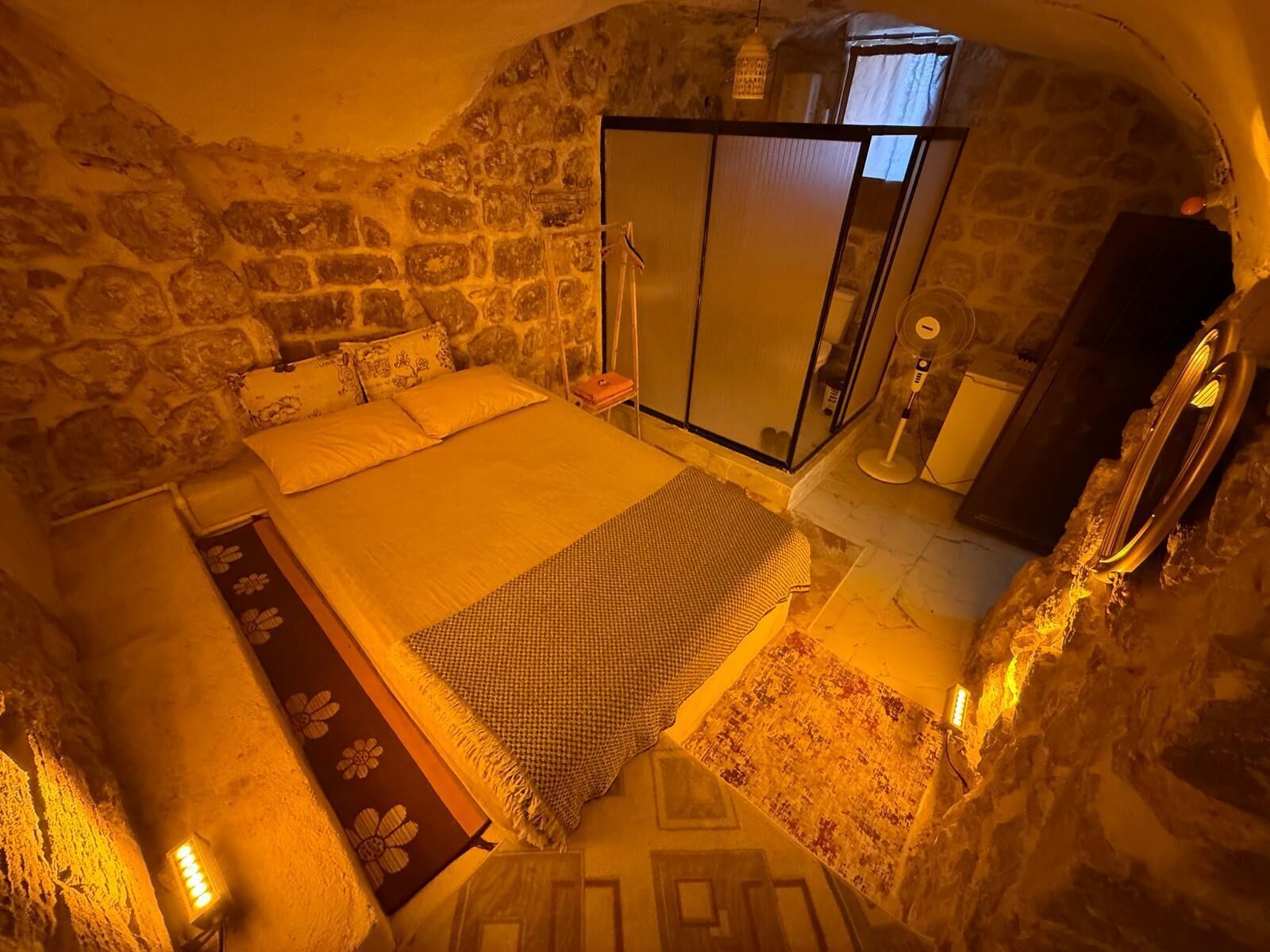 A stone-walled bedroom with a bed, a bedside table, a mirror, and a small window.