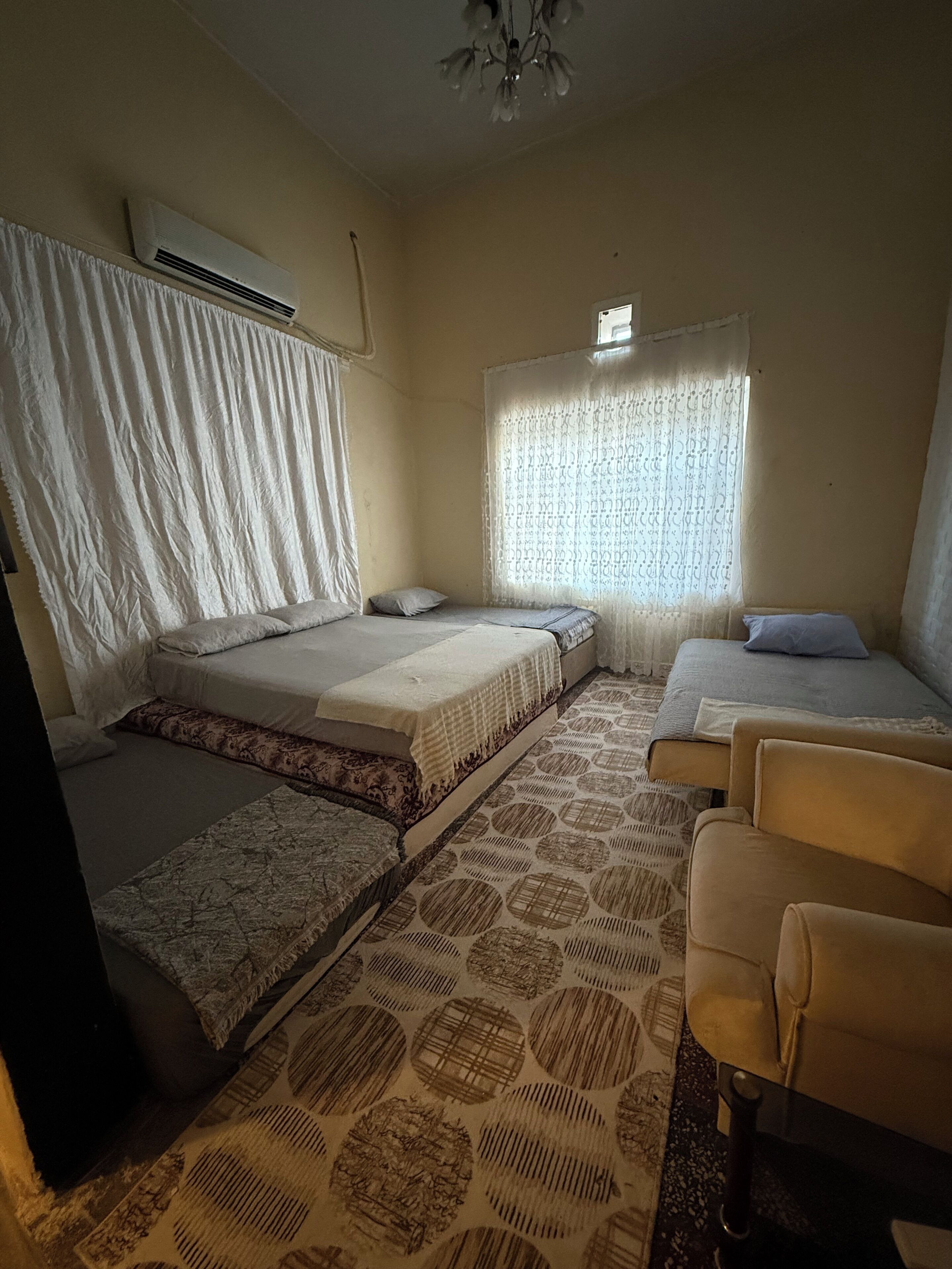 Una habitación con cama, sofá, alfombra estampada, ventana con cortinas y un equipo de aire acondicionado.