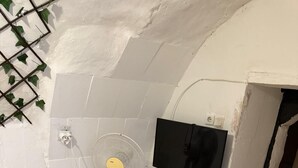 Una habitación pequeña y cerrada con piso estampado, un ventilador de pie, una mesita con un control remoto y un televisor montado.