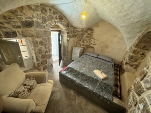 A cozy room with a stone wall, a bed with a marble-like cover, a small white cabinet, and a sofa with floral cushions.