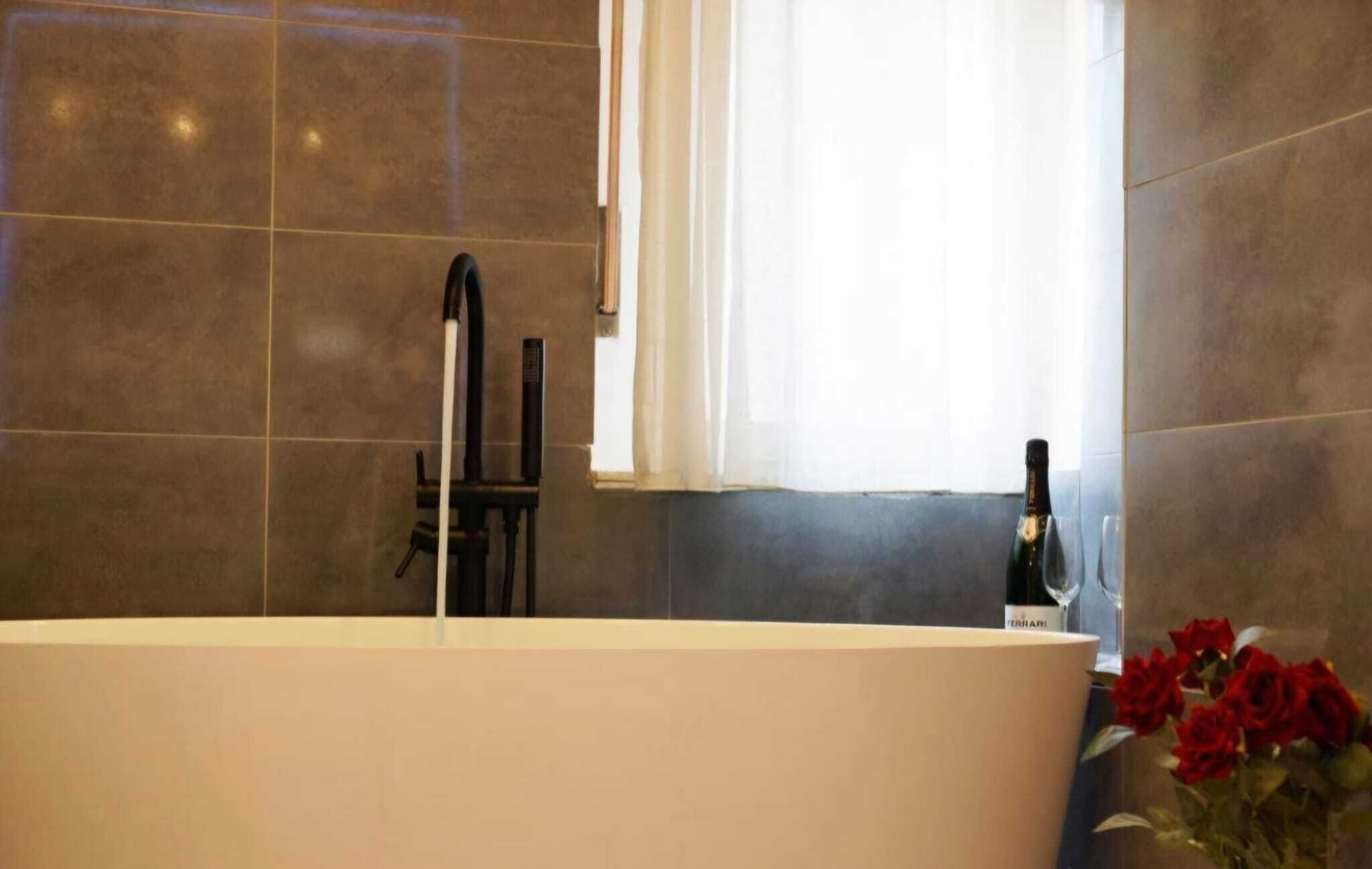 Ein Badezimmer mit einer freistehenden Badewanne, einem Fenster mit weißen Vorhängen und einer Weinflasche auf der Ablagefläche.