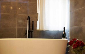 A bathroom with a freestanding bathtub, a window with white curtains, and a bottle of wine on the counter. - Alloggio in Pieno Centro a Trento  (Trento)