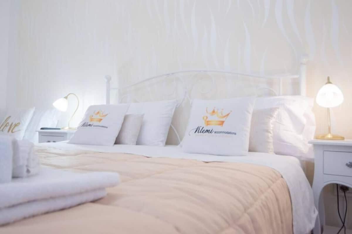 Een net opgemaakt bed met witte beddengoed en kussens, een bedlampje en een klein nachtkastje.