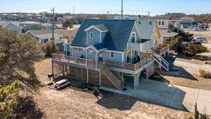 A two-story house with a balcony and a deck, surrounded by a sandy landscape and other houses. - The Wright Therapy, get your beach medicine here!  NEW LISTING w/ Hot Tub! (Kill Devil Hills)