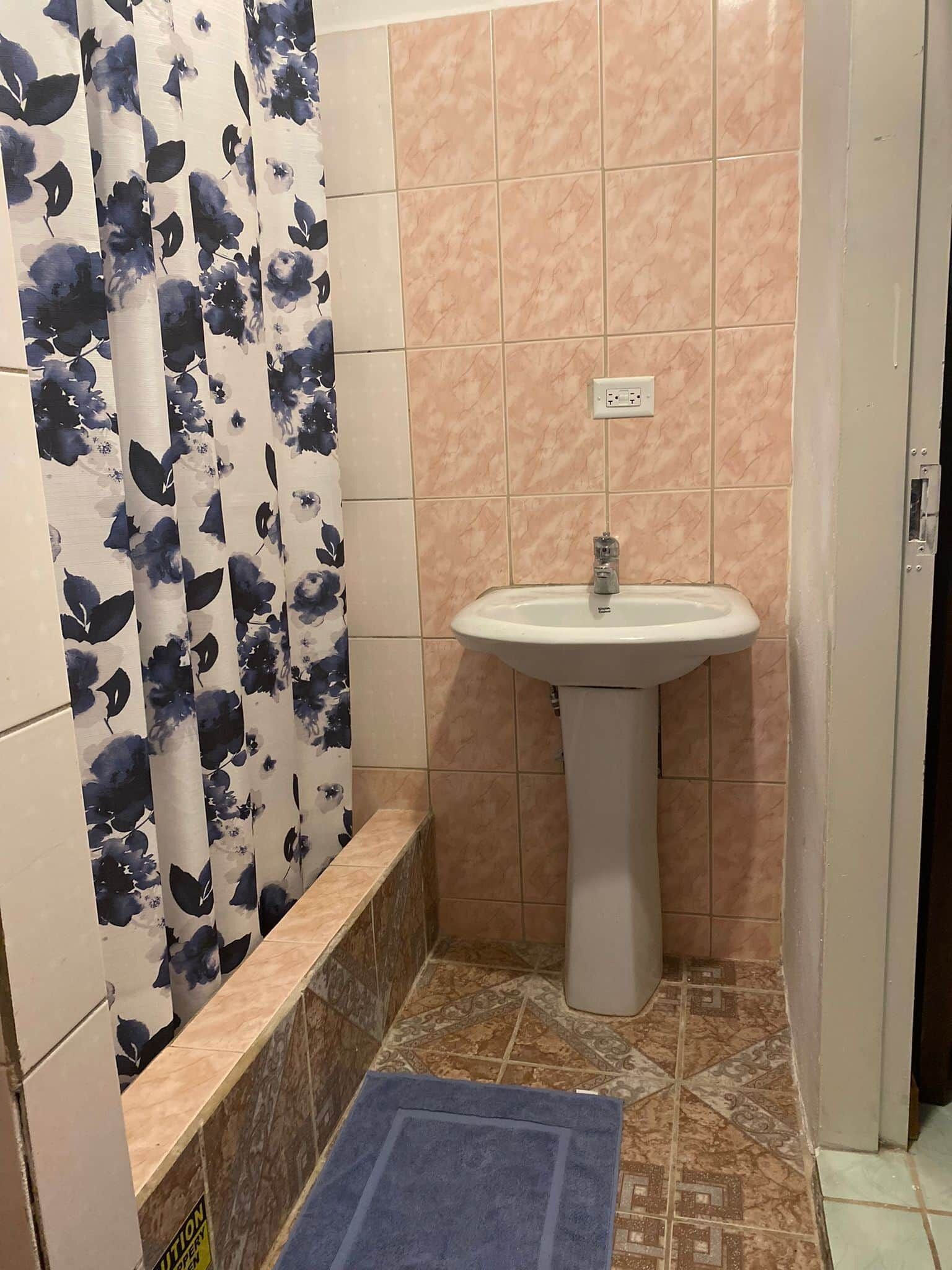 A bathroom with a sink, tiled walls, a shower curtain with a floral pattern, and a blue mat on the floor.