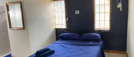 A bedroom with a bed, two windows with blinds, a mirror, and a wall-mounted air conditioner.