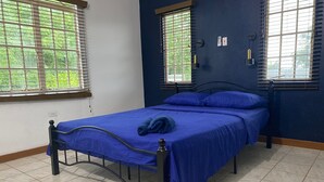 Un dormitorio con cama, dos ventanas con persianas, una lámpara de pared y un enchufe.