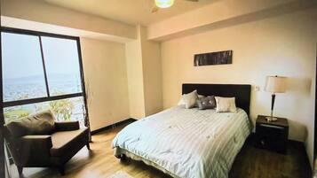 A bedroom with a large window, a bed with pillows, a chair, and a nightstand with a lamp.