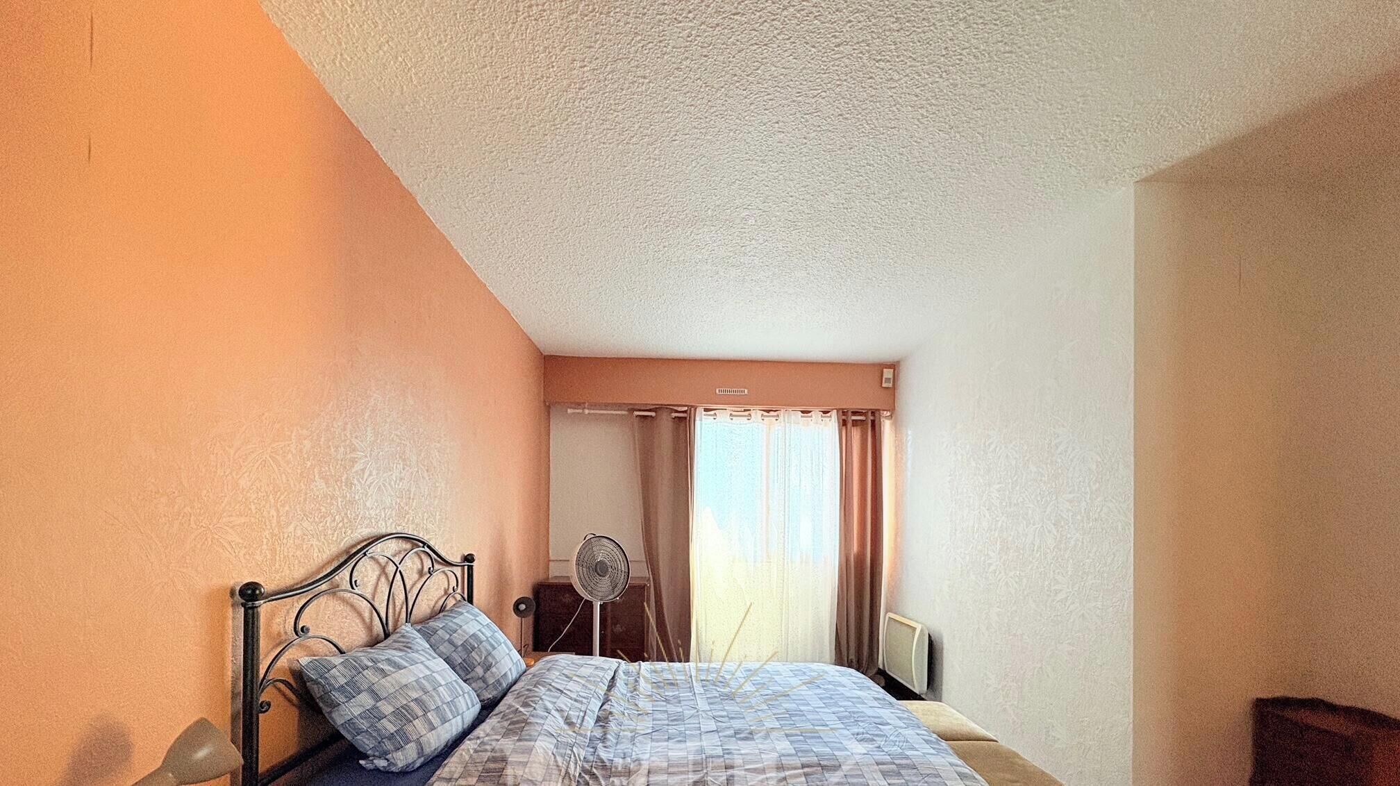 A bedroom with a bed, a bedside table, a fan, and a window with curtains.