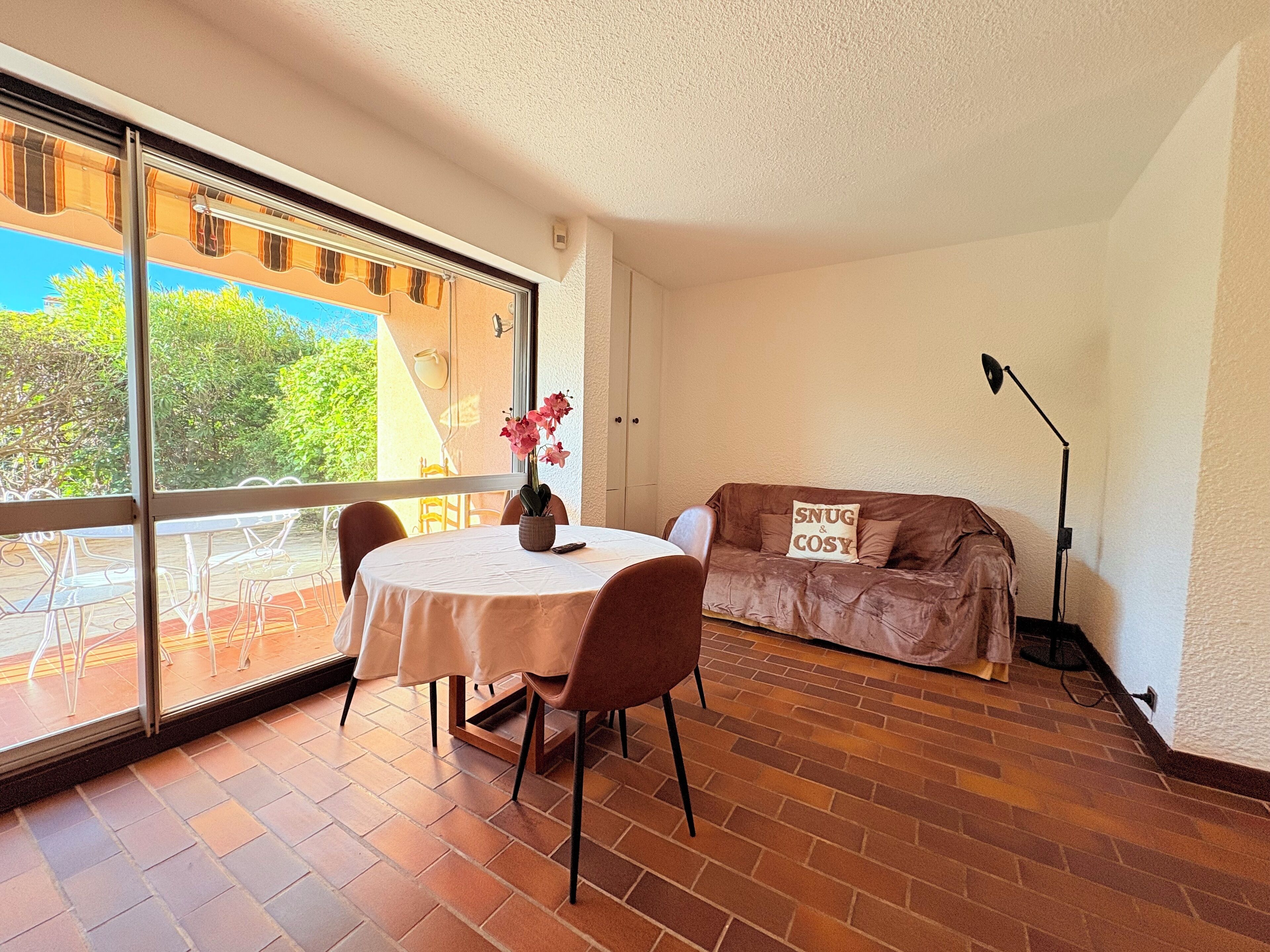 A room with a dining table and chairs, a sofa with a cushion, a floor lamp, and a large window with a view of greenery.