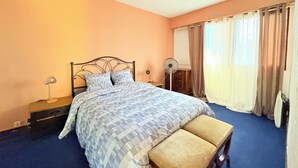Een slaapkamer met een bed, nachtkastje, lamp, ventilator en gordijnen.