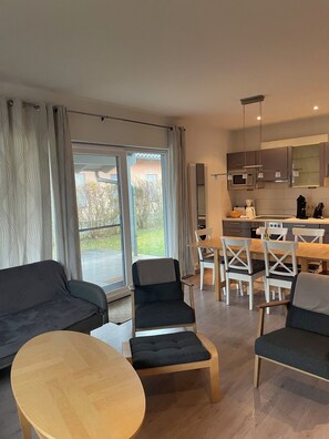 Ein modernes Wohnzimmer mit einer Couch, zwei Sesseln und einem Couchtisch. Es gibt eine Essecke mit Tisch und Stühlen. Das Zimmer verfügt über eine große Schiebeglastür, die auf einen Balkon führt.