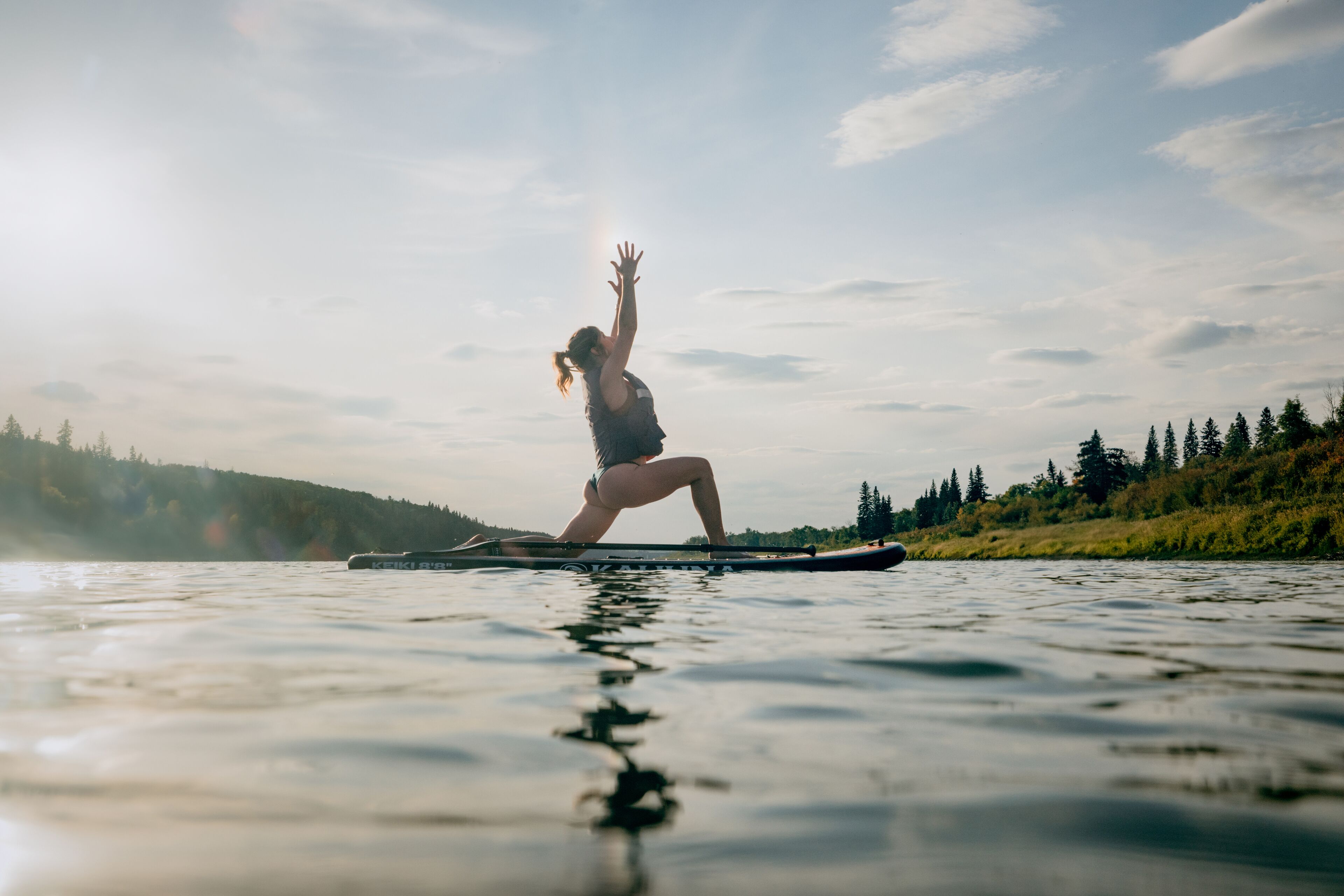A person practicing yoga on a paddleboard in a calm body of water with trees in the background.