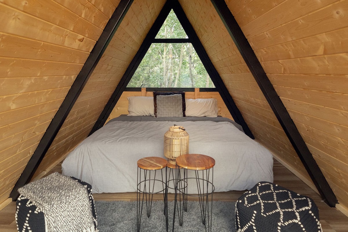 A cozy attic bedroom with a sloped ceiling, a bed with a grey blanket, a woven light fixture, and a view of trees through a window.