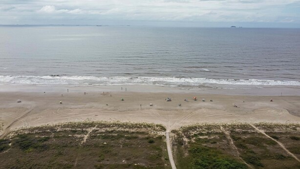 Vista aérea de una playa con orilla arenosa, algunas personas dispersas a lo largo de la playa y un mar tranquilo al fondo.