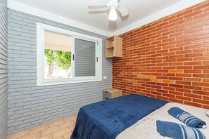 A bedroom with a grey brick wall, a wooden shelf, a bed with a blue bedspread, and a window with closed blinds.