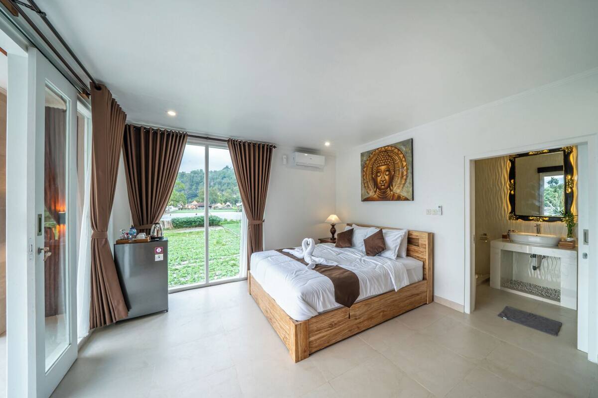 Een slaapkamer met een groot bed, een badkamer met een wastafel en spiegel, en uitzicht op groen buiten.