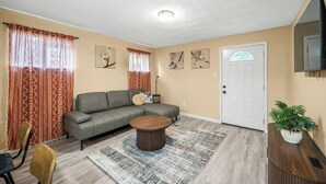 Un salon avec un canapé d’angle gris, une table basse ronde en bois, un tapis à motifs, une plante en pot et un téléviseur à écran plat fixé au mur.