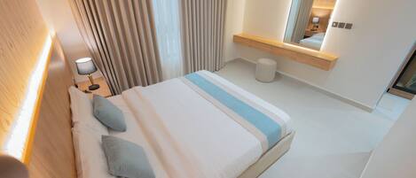 Una habitación de hotel moderna con una cama grande, una mesita de noche con lámpara, un espejo, un taburete pequeño y un cabecero de madera.