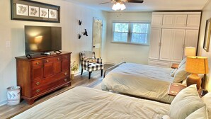 Une chambre avec deux lits, une commode, un téléviseur, un ventilateur de plafond et une fenêtre donnant sur des arbres.