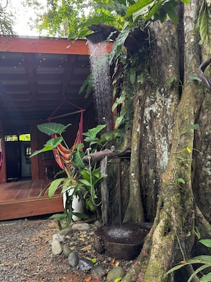 Un área de ducha al aire libre de estilo rústico con una estructura de madera, un árbol y un cabezal de ducha colgante.