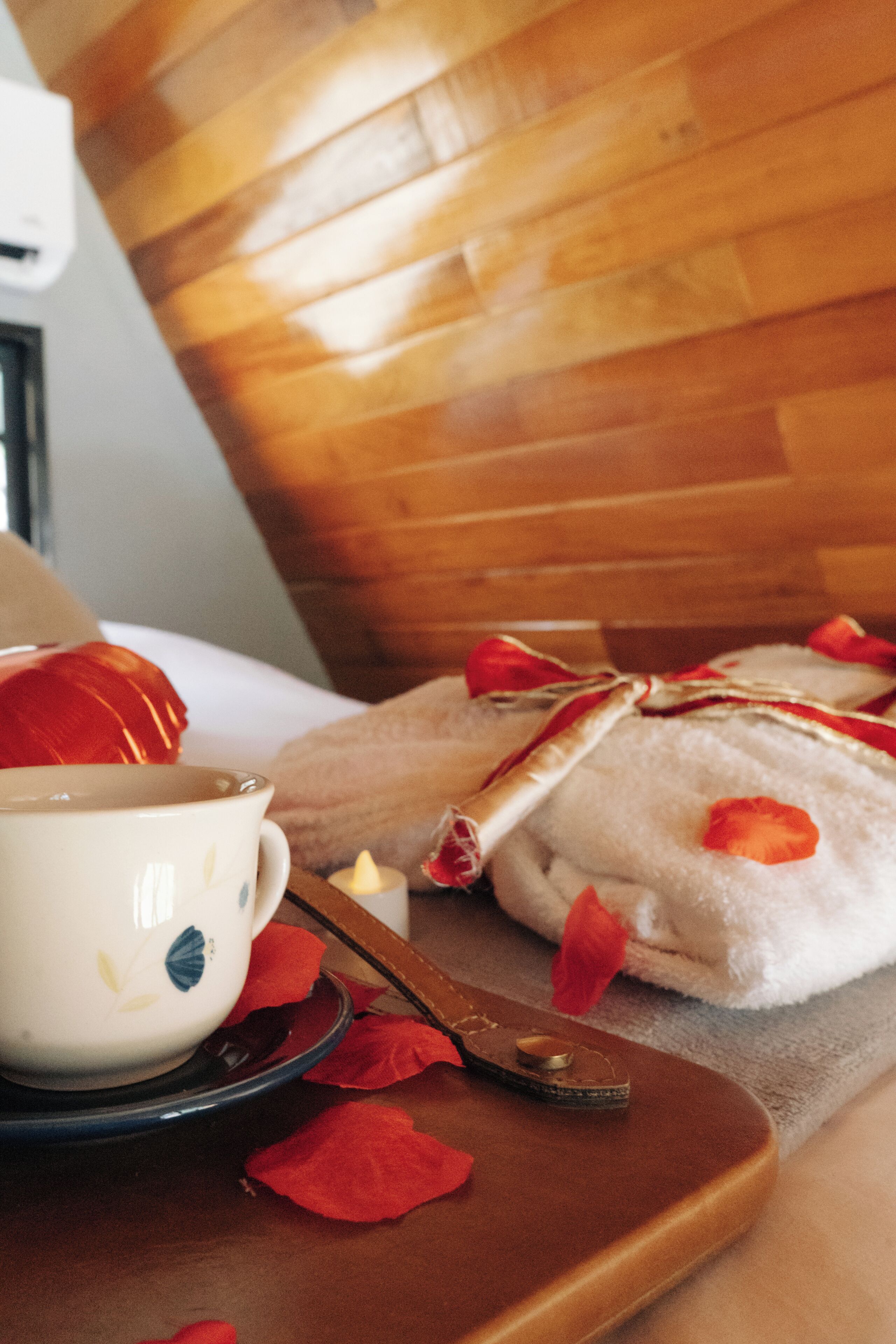 Une tête de lit en bois, une tasse blanche ornée de motifs floraux, un plateau marron avec une cuillère, et une serviette blanche parsemée de pétales de roses rouges.