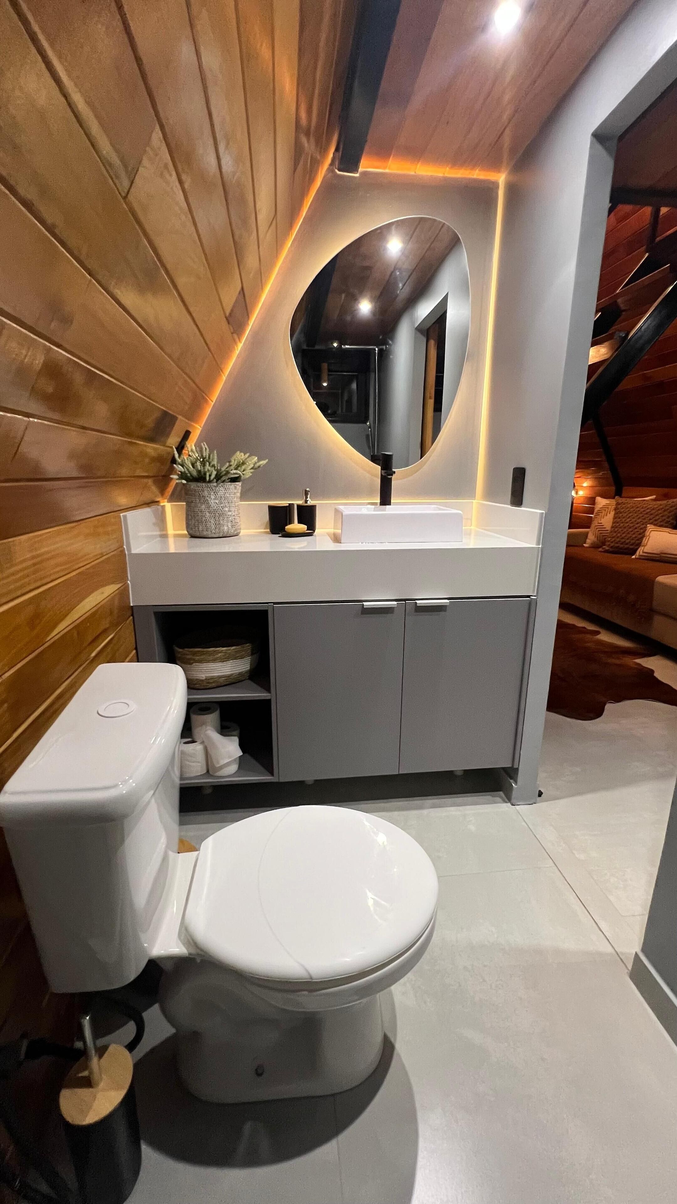Un baño moderno con pared de madera, un inodoro blanco, un lavamanos con espejo y un estante con artículos de aseo.