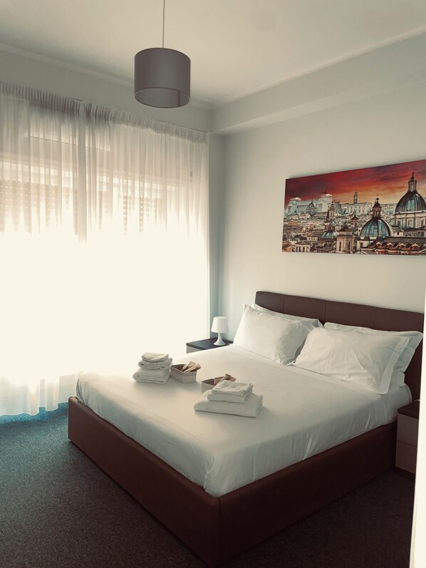A hotel room with a bed, a nightstand, a lamp, a painting of a cityscape, and sheer curtains. - A HOUSE SURROUNDED BY GREENERY, 5 MINUTES FROM ST. PETER, IN A QUIET SETTING (Roma)