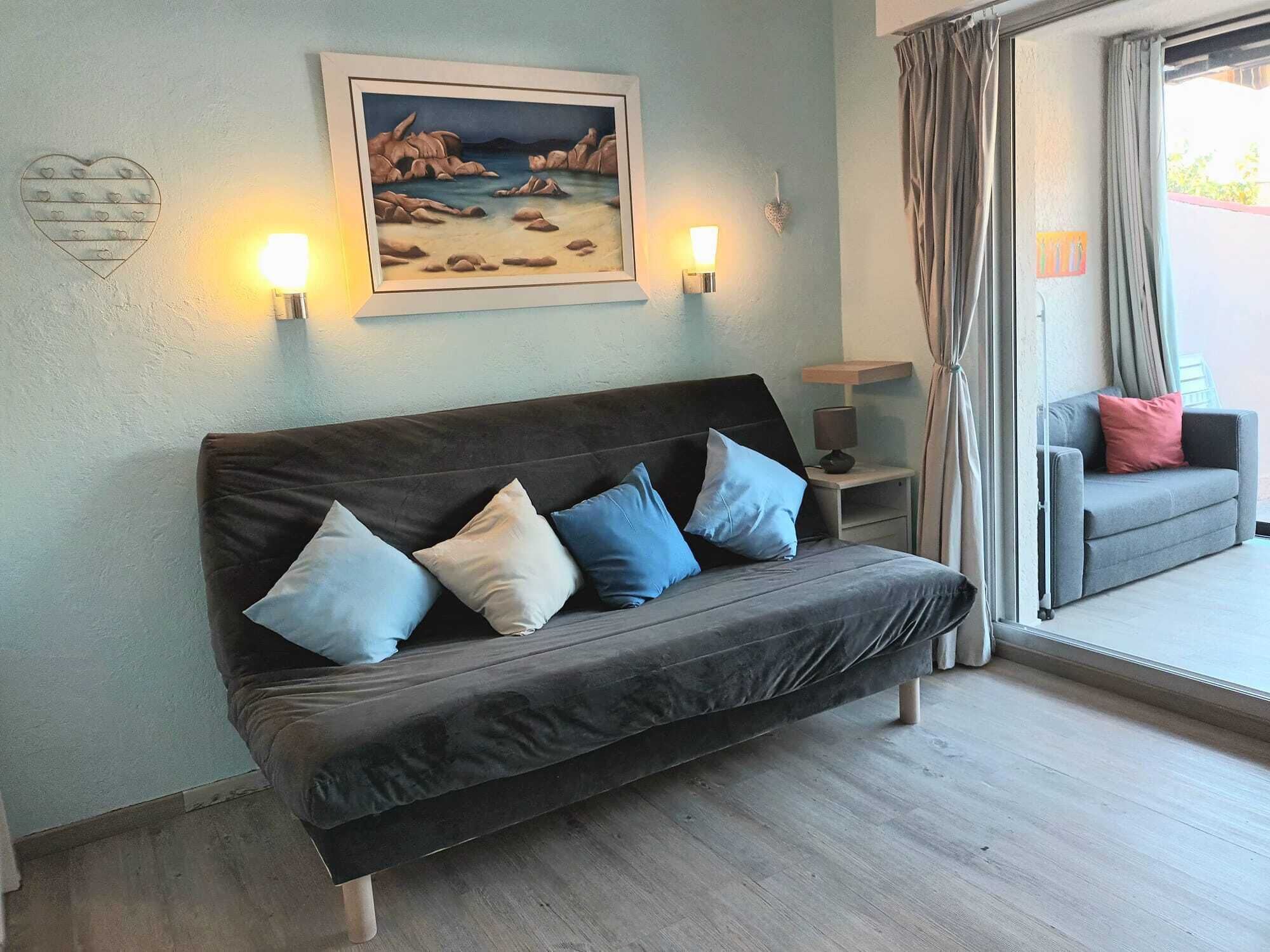 A dark grey sofa with four cushions in a room with light blue walls. There is a painting of a landscape above the sofa, two wall-mounted lamps on either side, and a small table with a lamp beside the sofa.