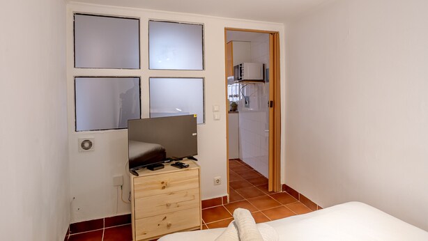 A bedroom with a bed, a dresser, a TV on a stand, and a kitchen visible through an open door. - Alfama Tiny by Homing (Lisbon)