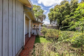 A covered porch with a brick pathway leading to a garden area with various plants and a wooden fence in the background.
