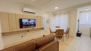 A modern living room with a flat-screen TV mounted on the wall, a brown leather sofa, a wooden dining table with chairs, and a window with sheer curtains. - Solarium Beach Club - Apartamento T1.307 (Itapoá)