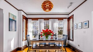 Una sala da pranzo con un tavolo in legno apparecchiato per sei persone, illuminata da due finestre con tende bianche, un banchetto in legno e un grande lampadario decorativo.