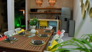 A dining area with a wooden table set for six, surrounded by white chairs with colorful cushions. A kitchen is visible in the background with a refrigerator and a microwave. - Casa Kolibrí - 8 Bedrooms + Pool + Outdoor Kitchen + Coworking (Mérida)