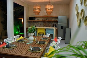 Een eetgedeelte met een houten tafel voor zes personen, omringd door witte stoelen met kleurrijke kussens. In de achtergrond is een keuken zichtbaar, met een koelkast en een magnetron.