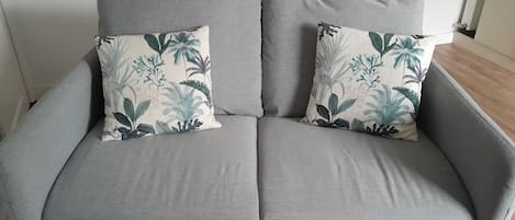 En grå soffa med två prydnadskuddar med blommönster och ett litet bord med vävda underlägg.
