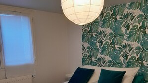 Et soveværelse med seng, blomstret sengetæppe, to puder, et vindue med persienner, en radiator og en væg med bladmotiv.