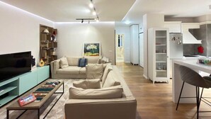 Un saló modern amb sofà, televisió i llibres, que connecta amb una cuina amb armaris blancs i zona de menjador.