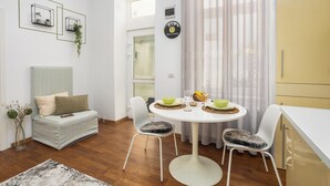 Et moderne spiseområde med et rundt bord, hvide stole og en hvid sofa. To vægmonterede hylder med planter, et ur og et vindue med gardiner.