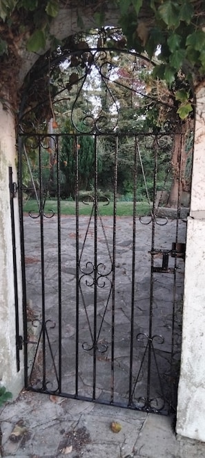 A wrought iron gate with intricate designs, leading to a paved area with a stone wall and greenery.