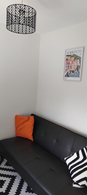 A black sofa with an orange and a black and white pillow, a patterned rug, a framed picture of a coastal town, and a geometric pendant light.