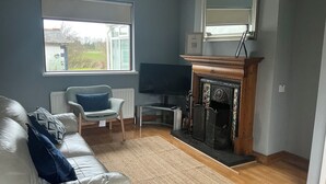 Een woonkamer met een witte bank, een blauw kussen, een open haard, een televisie en een groot raam.