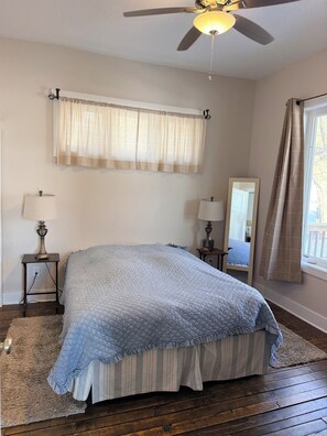 Un dormitorio con cama, ventilador de techo, ventana con cortinas, mesita de noche con lámpara y espejo.