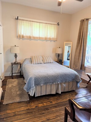 Un dormitorio con una cama, dos mesitas de noche, un espejo, una ventana con cortinas y piso de madera.