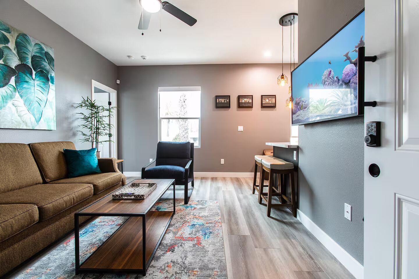 A living room with a brown sofa, a black armchair, a wooden coffee table, and a colorful rug. There is a window with blinds, a ceiling fan, and a mounted TV on the wall.